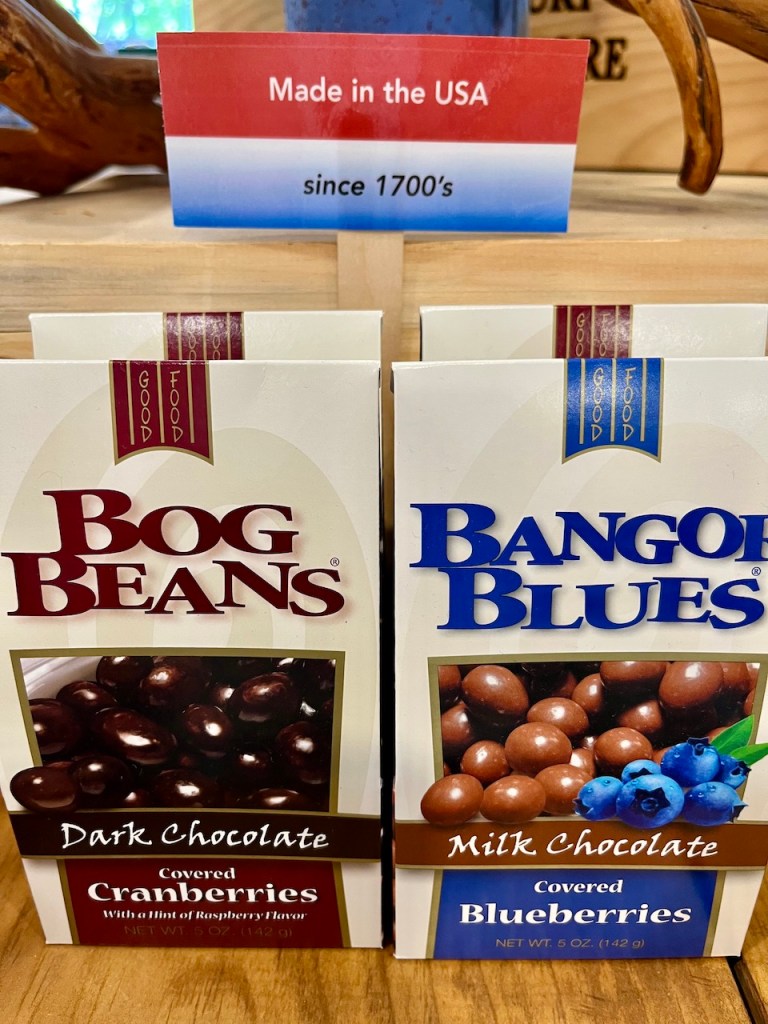 Two boxes of chocolate-covered snacks on a wooden display. The left box is labeled 'Bog Beans Dark Chocolate Covered Cranberries' and the right box is labeled 'Bangor Blues Milk Chocolate Covered Blueberries.' A sign above reads 'Made in the USA since 1700's.'