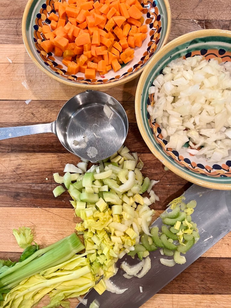 Chopped celery, carrots, and onions arranged on a wooden cutting board next to a measuring cup.