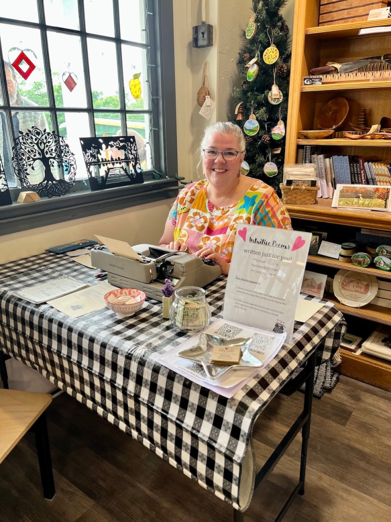 A woman sitting at a table with a typewriter, colorful shirt, and a decorative store background, promoting intuitive poetry services.