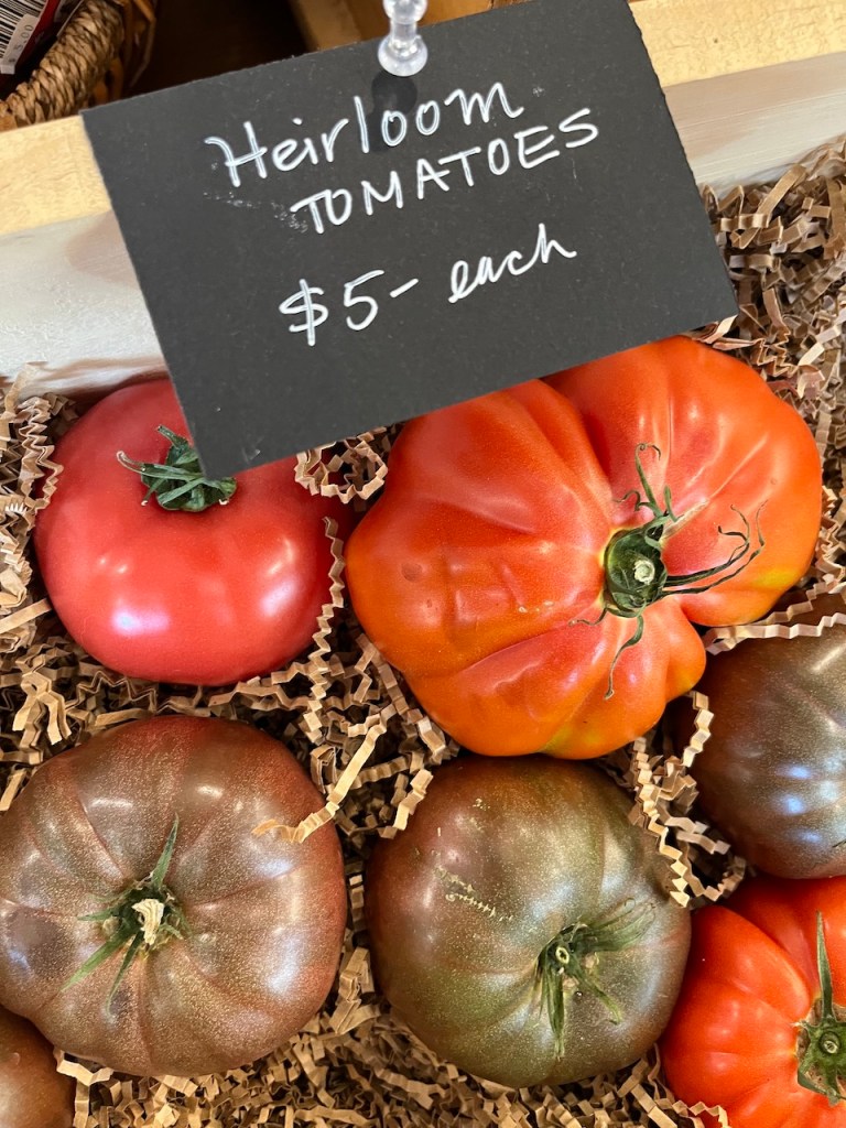 A variety of heirloom tomatoes with a sign indicating their price of $5 each, displayed in a wooden crate filled with shredded paper.