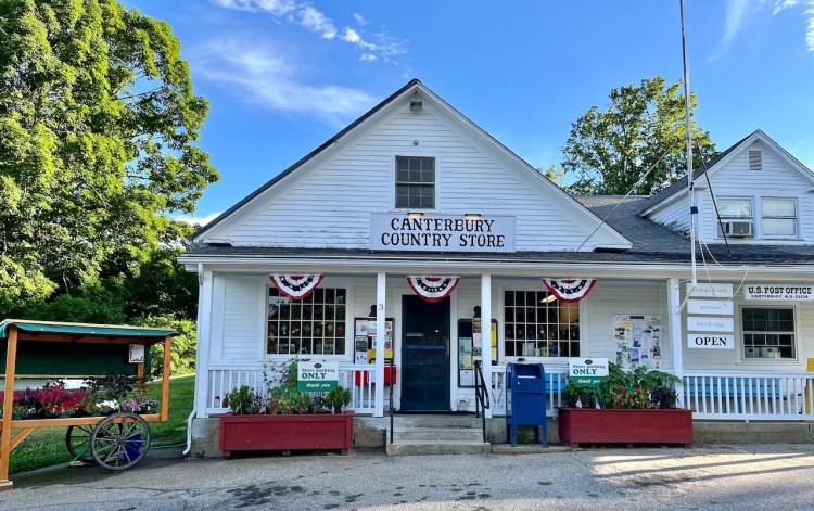 Exterior view of the Canterbury Country Store, showcasing white siding, a sloped roof, and festive decorations including bunting in red, white, and blue. The store features a front porch with plants and an open sign, set against a backdrop of green trees.