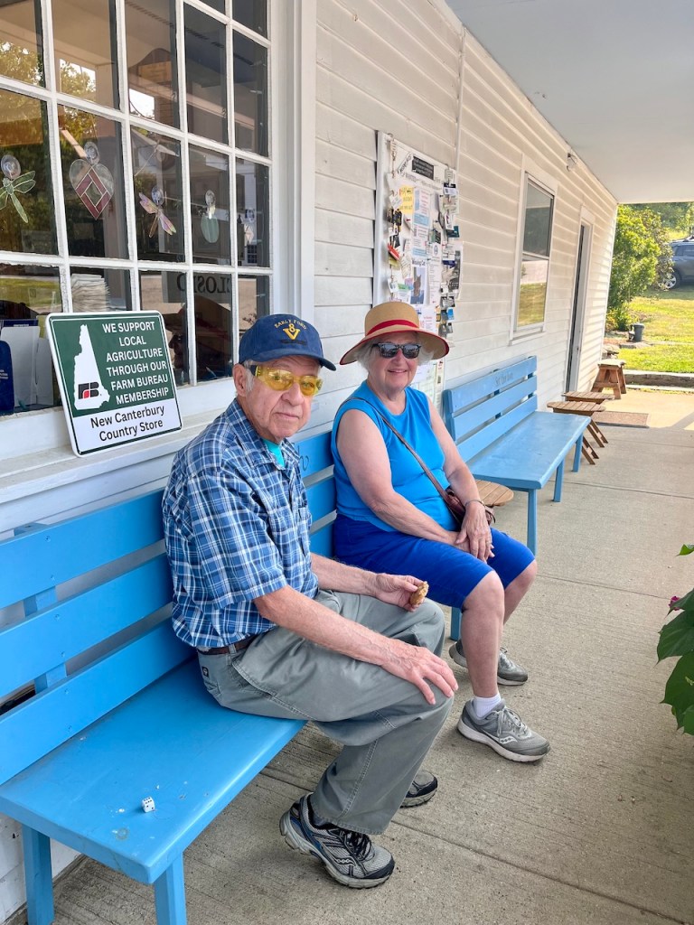 An elderly man in a plaid shirt and cap sits on a blue bench next to an elderly woman wearing a blue shirt and sun hat, both relaxed outside a store.