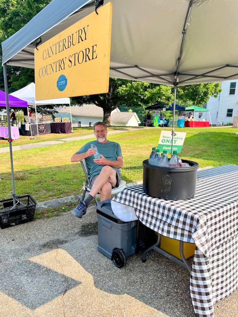A man sitting under a tent with a sign for Canterbury Country Store, holding a bottle of water, at a summer fair with colorful tents in the background.