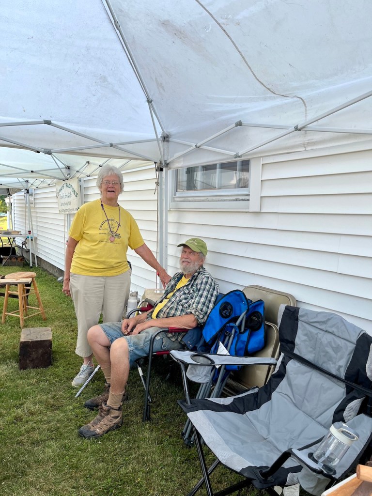 A smiling elderly woman in a yellow shirt stands beside an older man sitting in a chair under a canopy, with outdoor furniture and a shed in the background.
