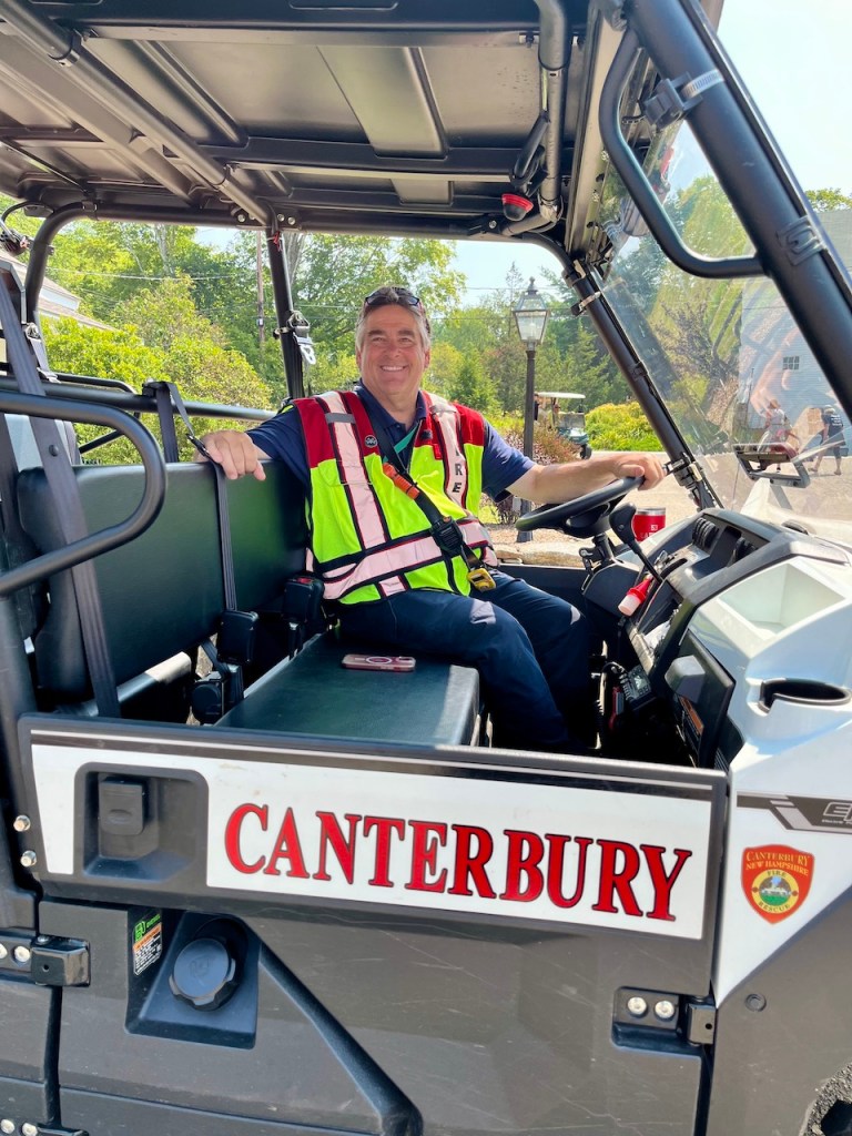 A smiling man sitting in a vehicle, wearing a safety vest with 'Canterbury' displayed on the side.