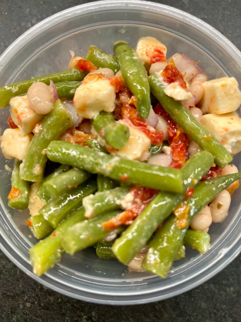 A colorful bowl of green bean salad featuring green beans, cheese, sun-dried tomatoes, and white beans, garnished in a clear container.