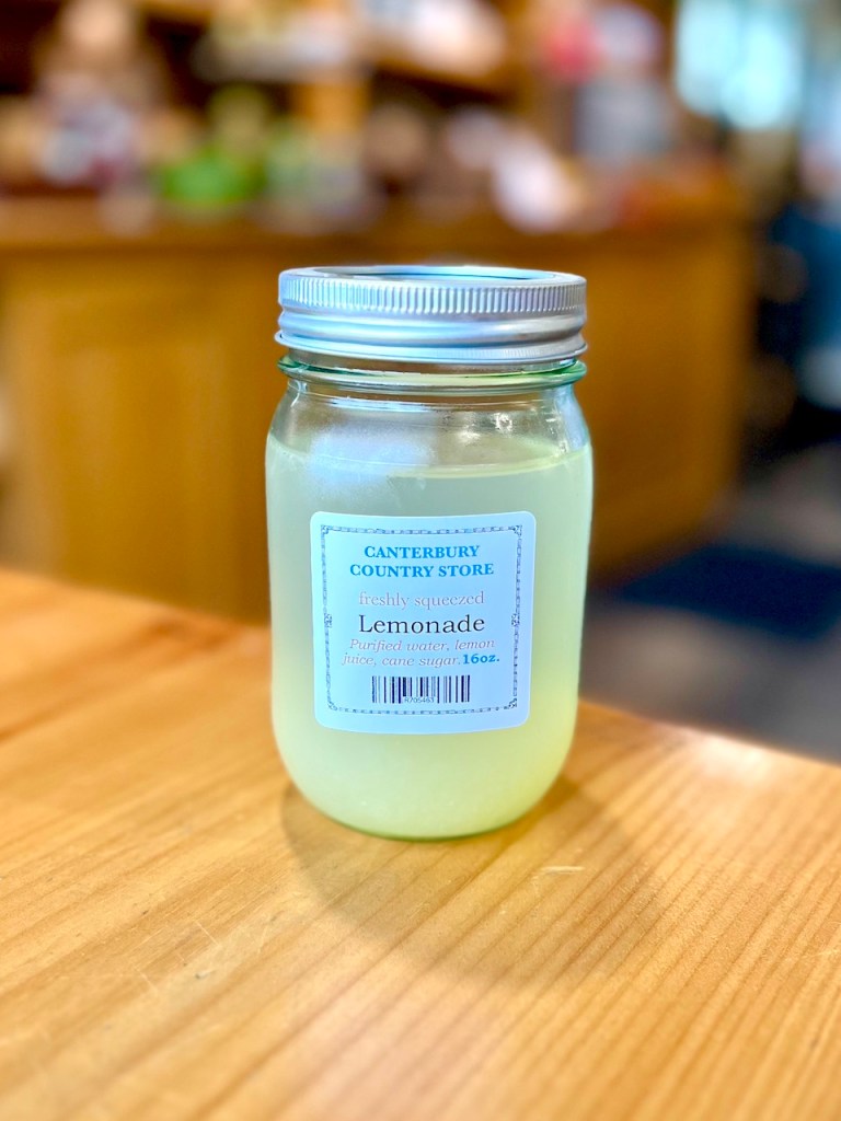 A jar of freshly squeezed lemonade with a label indicating it contains purified water, lemon juice, and cane sugar, placed on a wooden table in a store.