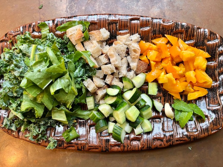 A wooden platter filled with chopped fresh ingredients for a summer soup, including kale, spinach, diced sourdough bread, sliced cucumbers, yellow tomatoes, mint leaves, and a clove of garlic.