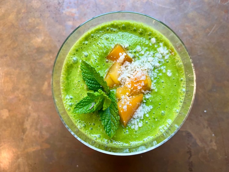 A refreshing summer green soup garnished with fresh mint leaves and diced peaches, served in a clear glass bowl.