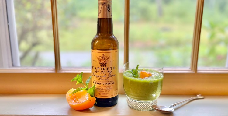A bottle of sherry vinegar labeled 'Capirete' next to a glass of green soup garnished with mint and a slice of apricot, resting on a windowsill with a scenic background.