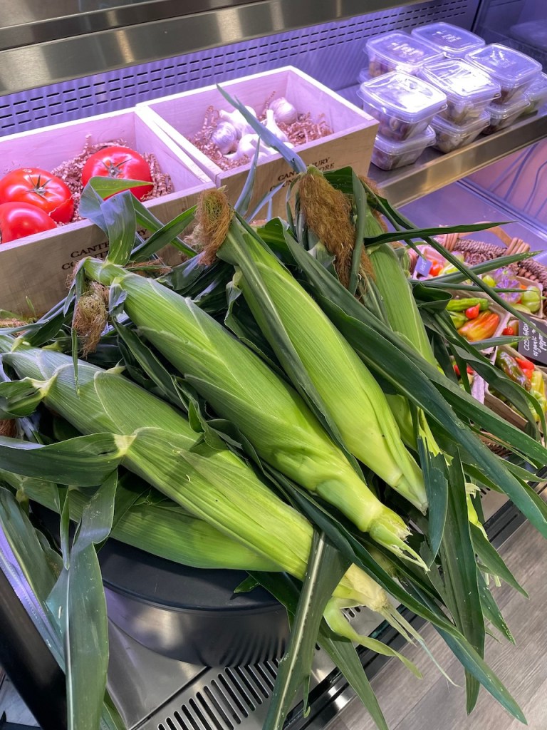 Fresh corn on the cob displayed in a grocery setting, surrounded by ripe tomatoes and garlic in wooden boxes.