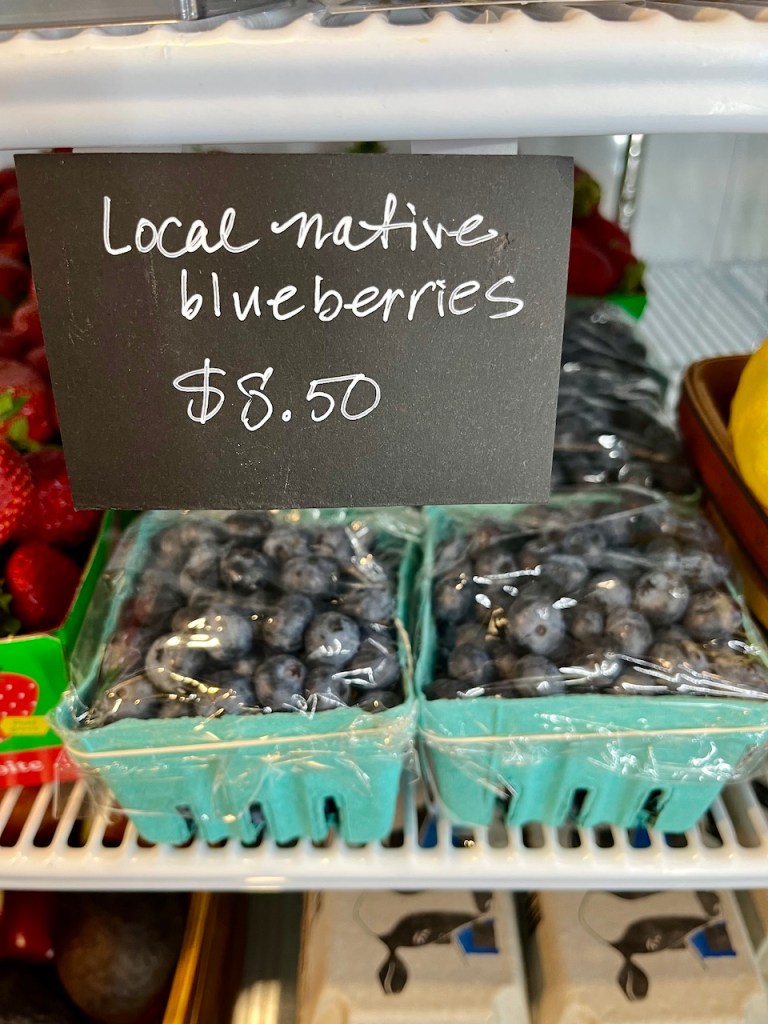A display of local native blueberries in baskets, with a price tag of $8.50, placed next to strawberries and other produce.