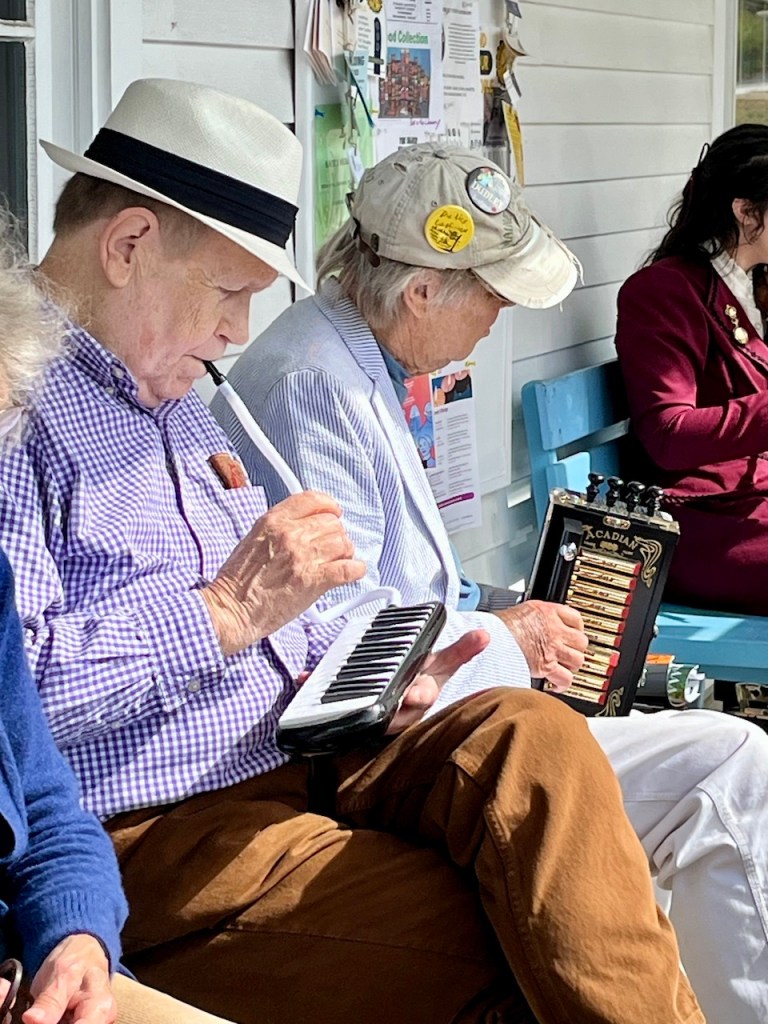 Two elderly musicians playing instruments on a porch. One man is playing a melodeon while the other plays a small keyboard recorder. They are sitting next to each other, engaged in their performance.