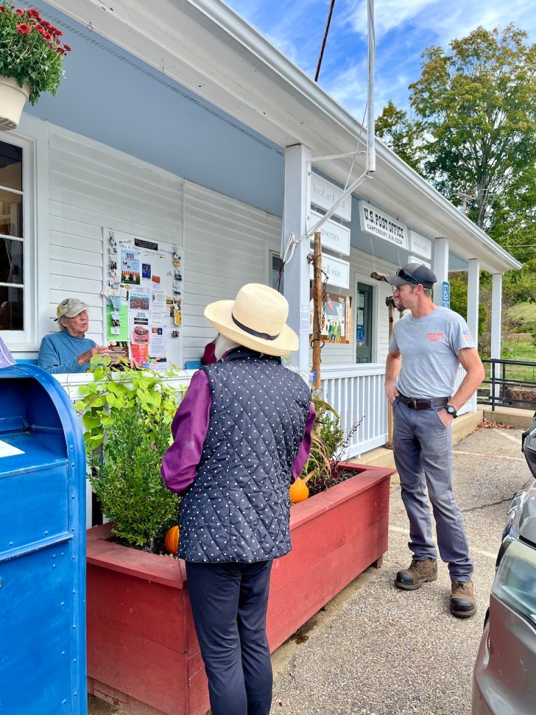A woman in a wide-brimmed hat and a quilted vest is talking to a postal worker at a window of a small post office. Another man stands nearby, observing the interaction. The post office features a bulletin board with various flyers and is surrounded by greenery and pumpkins.