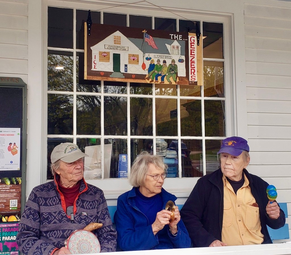 Three elderly individuals sitting together on a porch, each holding a musical instrument. A sign above them reads 'The Geezers.'