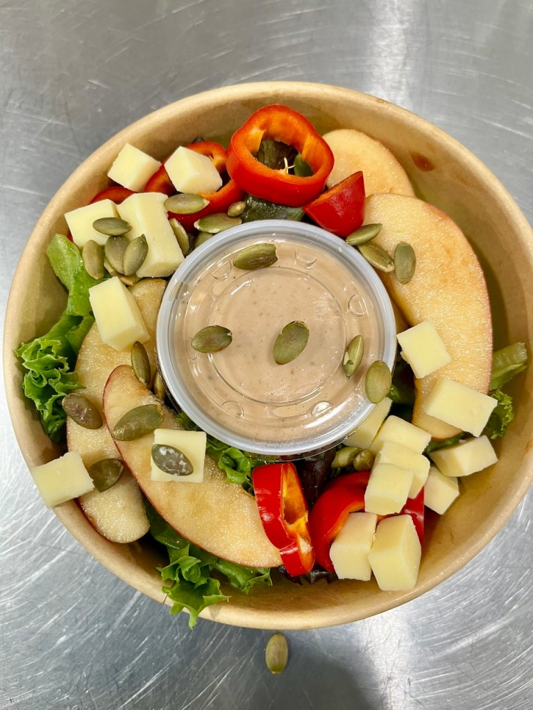 A bowl of fresh salad with lettuce, sliced apples, diced cheese, red bell peppers, and pumpkin seeds, topped with a container of dressing.