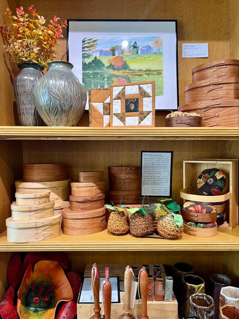A wooden shelf displaying various handcrafted items, including decorative boxes, ceramics, and art. There are two vases with dried flowers and a framed painting of a rural landscape in the background.