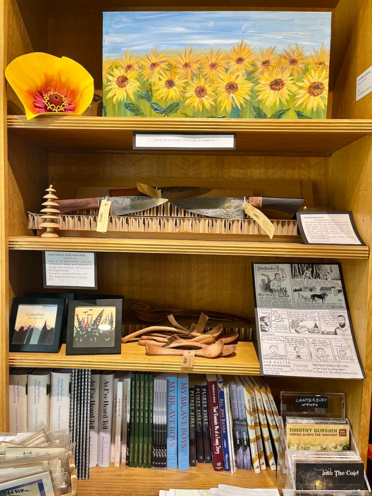 A wooden shelf displaying various handcrafted items including a sunflower painting, kitchen knives, wooden utensils, framed artwork, and a collection of books.