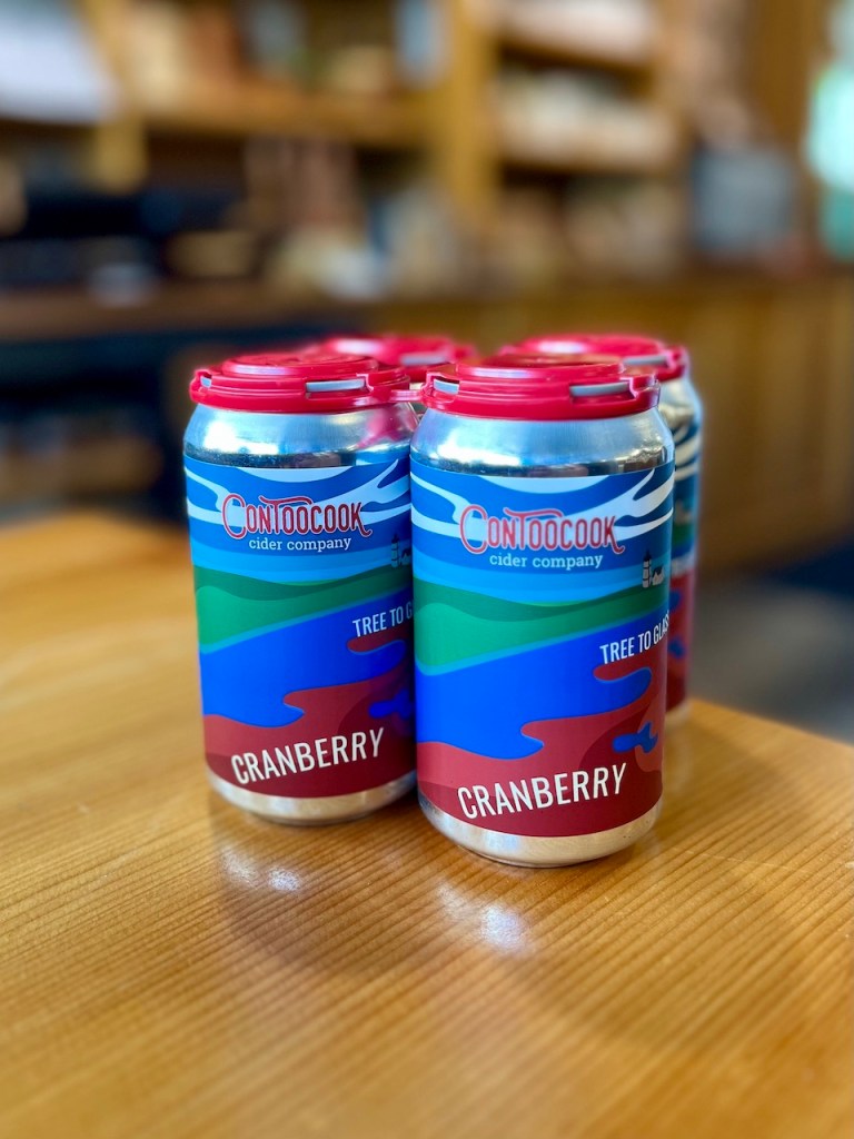 Six cans of Contoocook cider, displayed on a wooden table, labeled 'Tree to Glass' and 'Cranberry'.