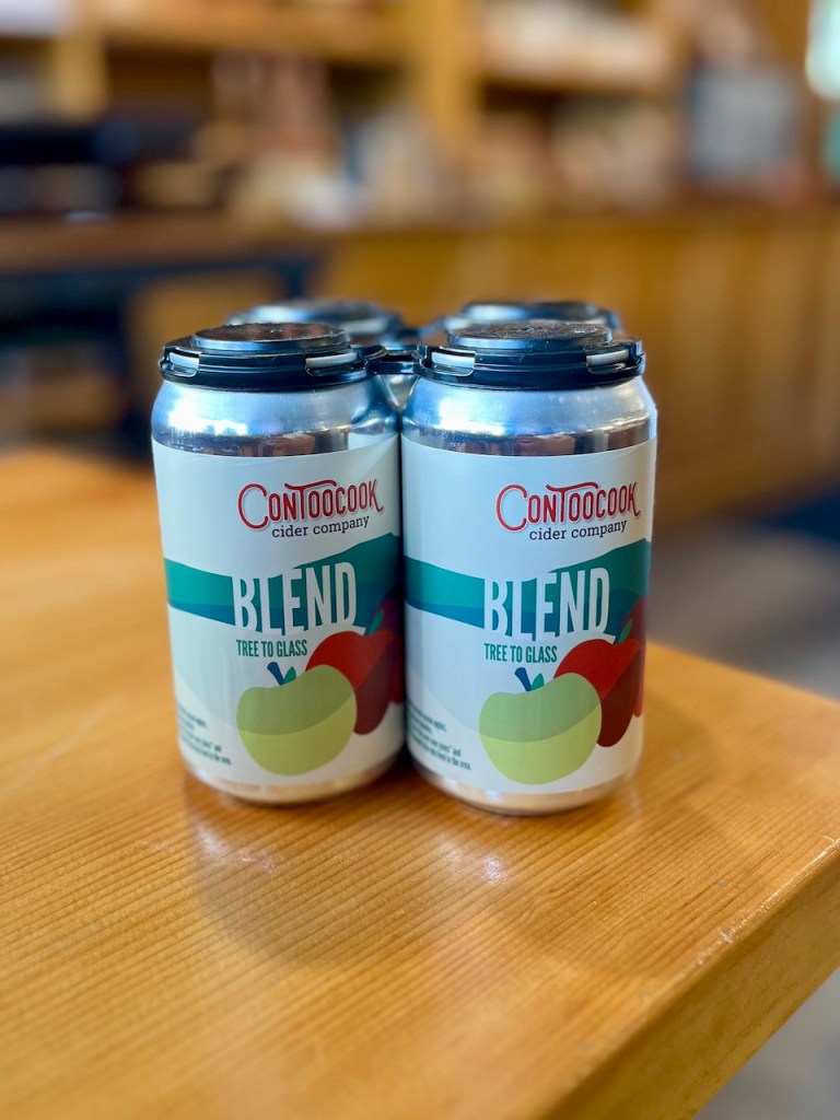 Four cans of ConToocook cider blend placed on a wooden table, with a blurred background.