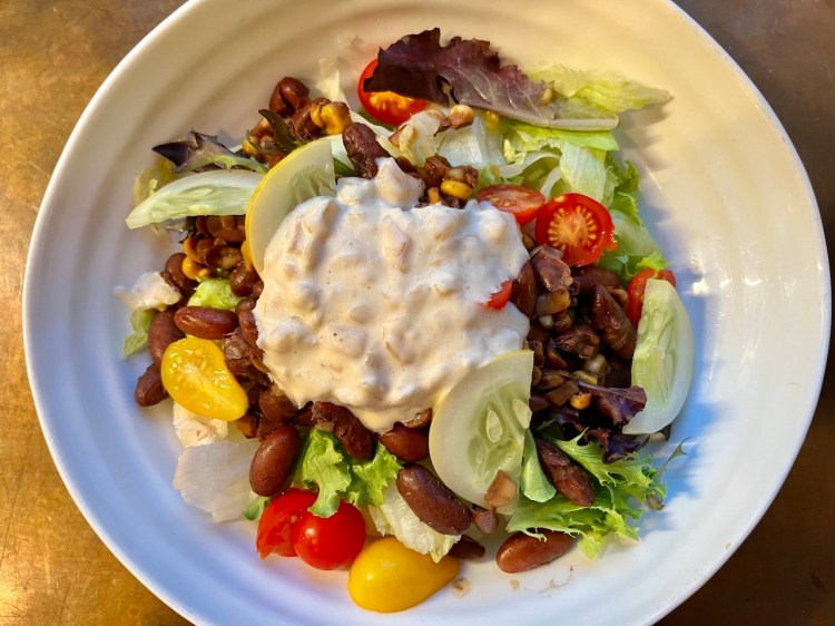 A colorful salad featuring a mix of fresh greens, heirloom beans, corn, cherry tomatoes, and slices of cucumber, topped with a creamy aioli dressing.