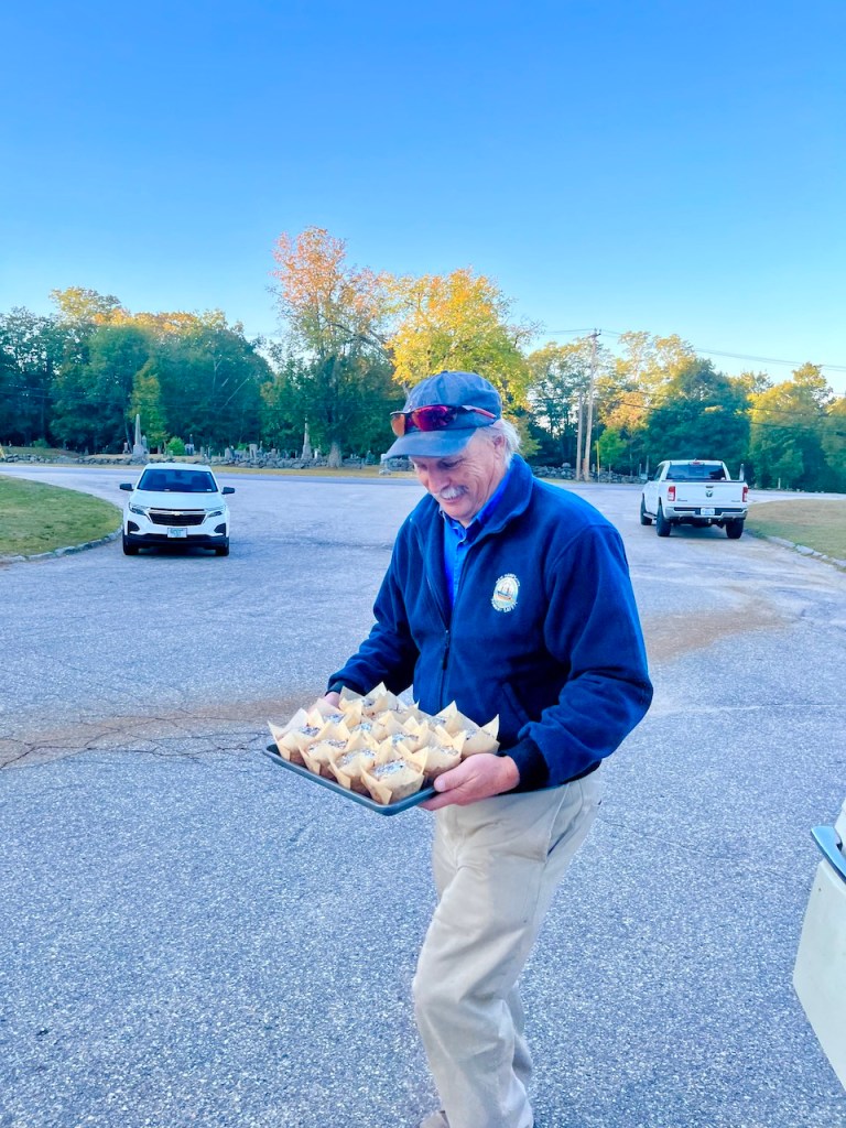 A man smiling while carrying a tray of baked goods outdoors, with trees in the background and parked vehicles in a parking lot.