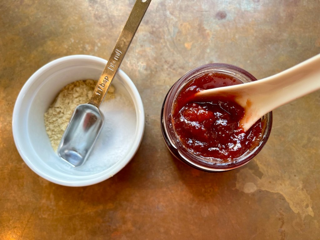 A measuring spoon next to a small bowl containing dried mustard and kosher salt, alongside a jar of cherry jam with a spoon resting on the edge.