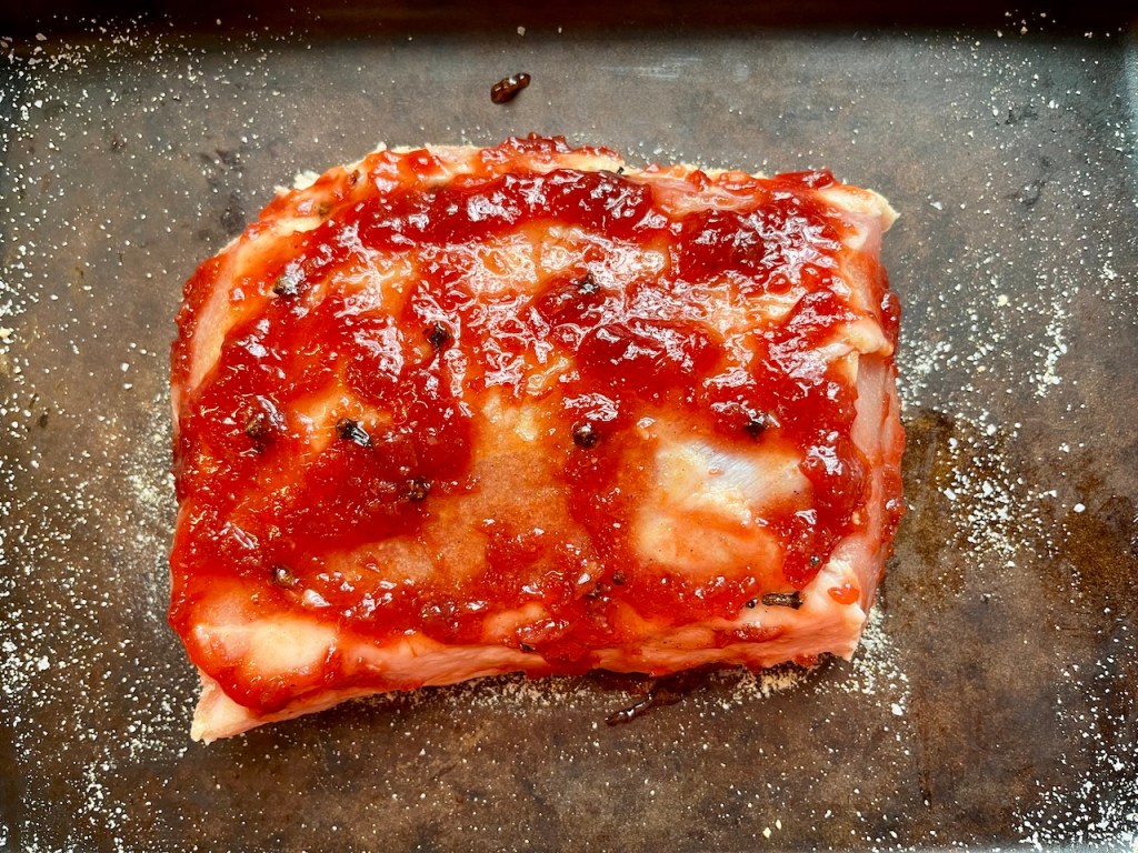 A cooked pork loin roast topped with a layer of cherry jam glaze, resting on a baking sheet.