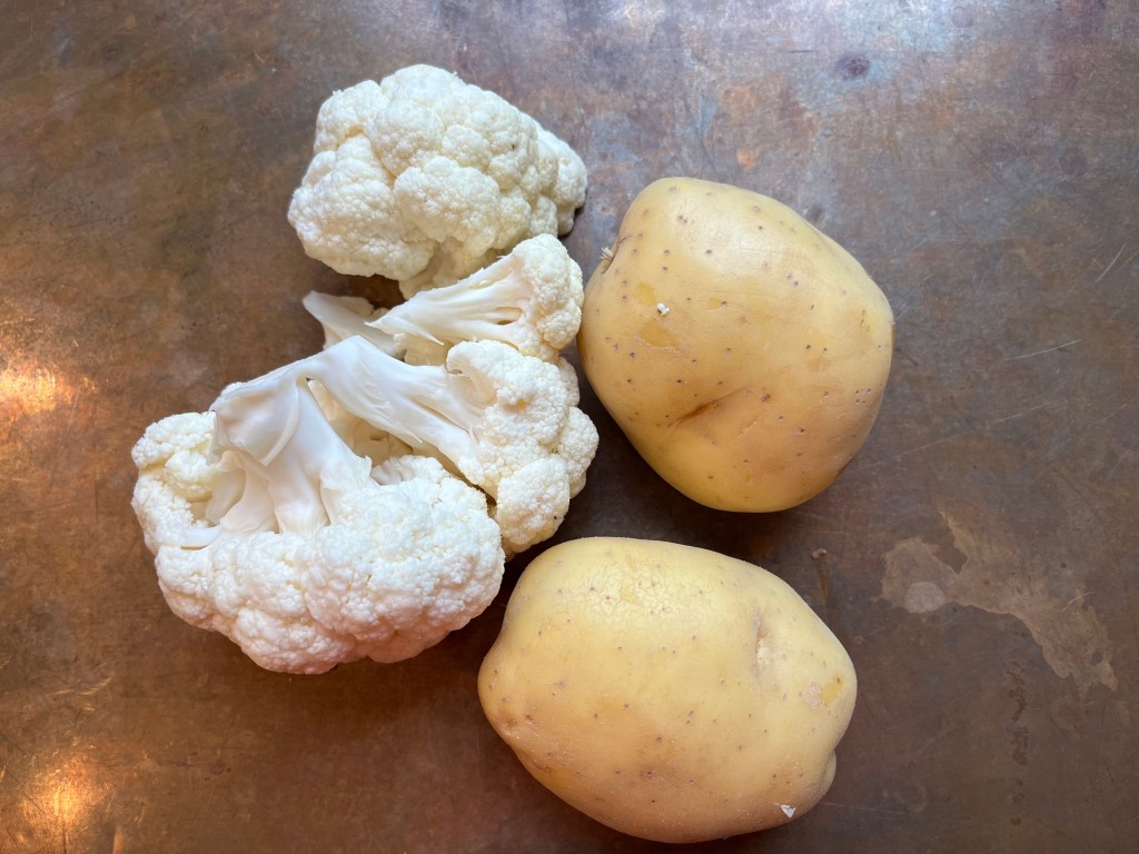 A head of cauliflower and two Yukon gold potatoes on a rustic surface.