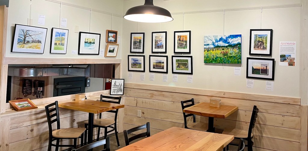 An interior view of a cozy art gallery featuring wooden tables and black chairs. The walls display various framed artworks and paintings, with a glimpse of a kitchen area in the background.