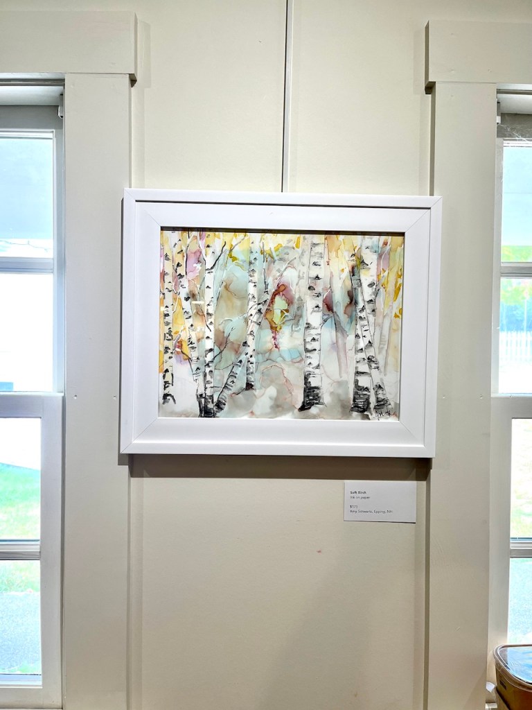 A framed watercolor painting depicting birch trees with autumn-colored leaves, displayed on a wall near a window.