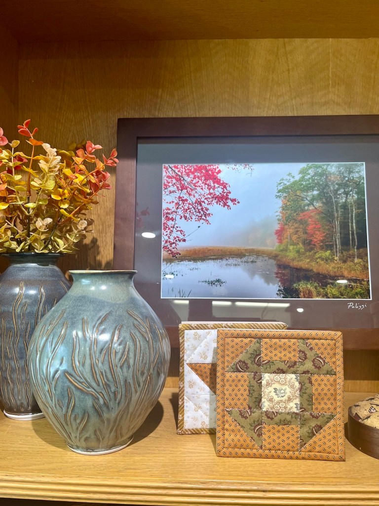 A decorative wooden shelf displaying a blue ceramic vase with a textured design, a framed autumn landscape photograph with vibrant red leaves, and a quilted wall hanging featuring a star pattern.