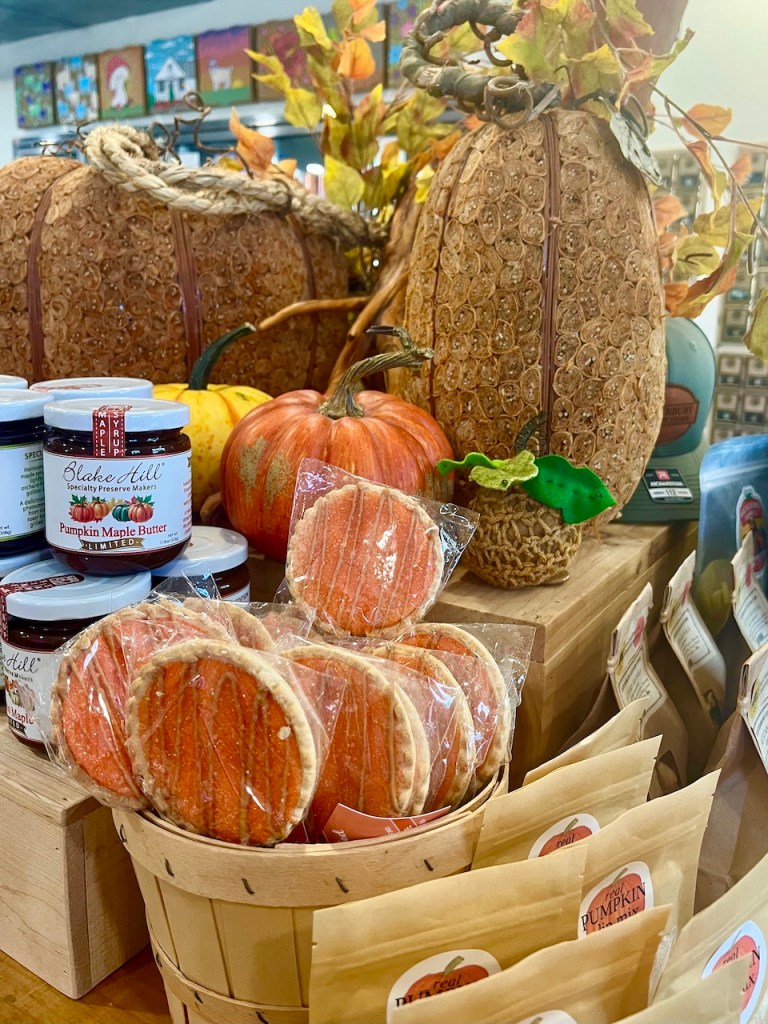 A display of autumn-themed decor featuring pumpkins, leaves, and seasonal treats including pumpkin cookies and jars of preserves in a country store setting.
