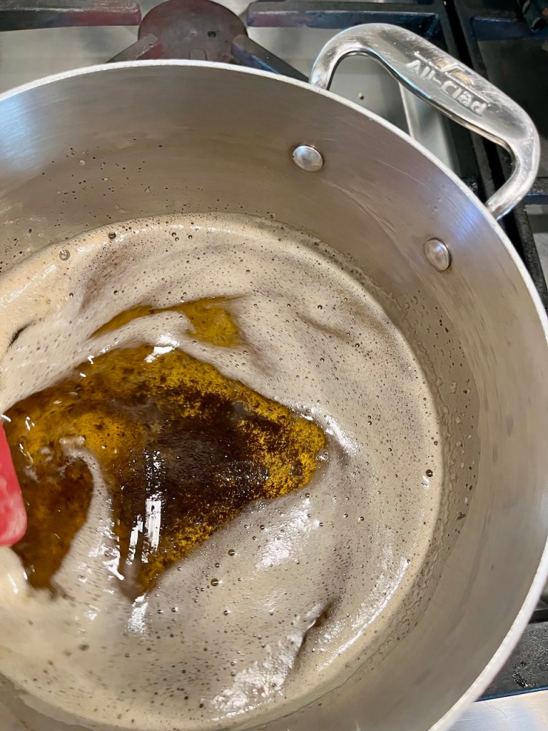 A close-up view of a pot on the stove with boiling butter, showing a foamy, golden liquid and a spatula stirring it.