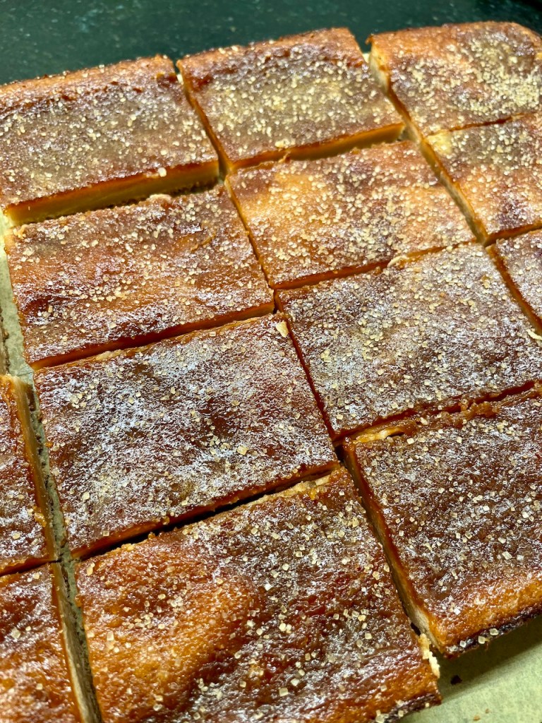 A close-up view of a freshly baked, golden-brown cake, cut into square pieces and topped with sparkling sugar.