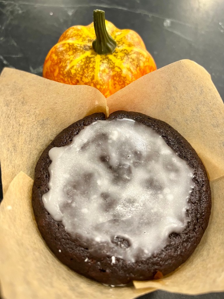 A close-up of a glazed chocolate cookie sitting on a piece of parchment paper, with a small, decorative pumpkin in the background.