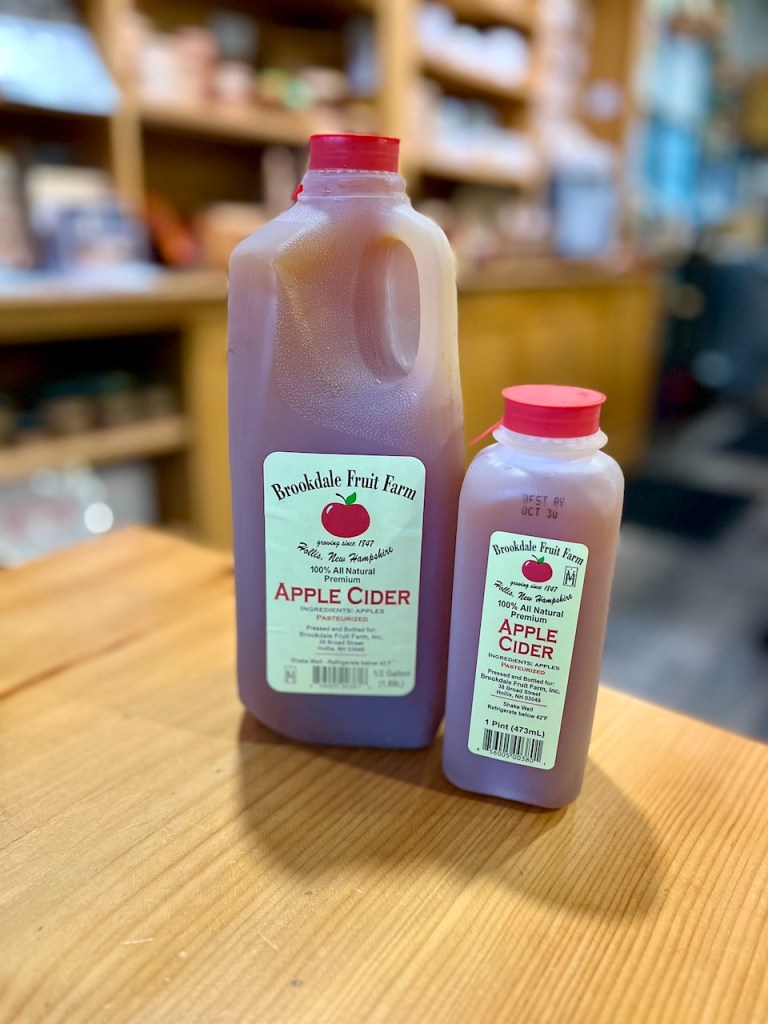 Two bottles of Brookdale Fruit Farm apple cider on a wooden table, one large and one small, with labels indicating 100% all-natural ingredients.