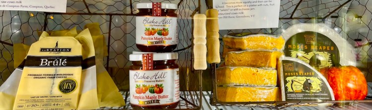 A shelf display featuring jars of pumpkin maple butter and a selection of artisanal cheeses, alongside wooden cheese utensils and a sign with information about the products.