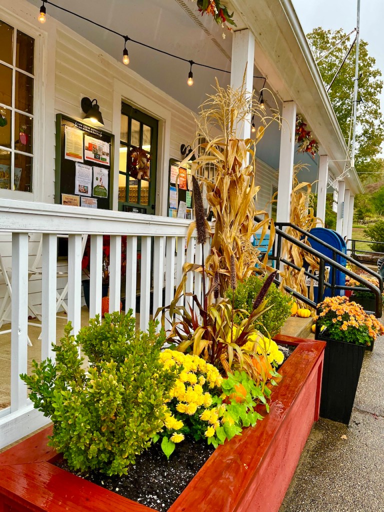 A view of a cozy storefront decorated for autumn, featuring corn stalks, flowering plants, and cozy lighting.