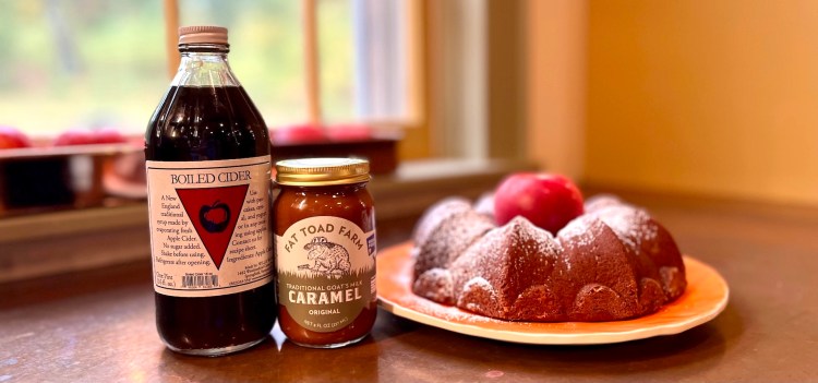 A bottle of boiled cider, a jar of caramel, and a bundt cake dusted with powdered sugar on a plate, accompanied by a red apple.