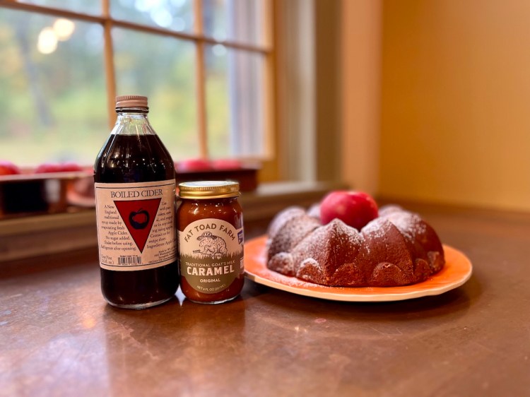 A bottle of boiled cider, a jar of goat milk caramel, and a bundt cake are arranged on a table with a window in the background.