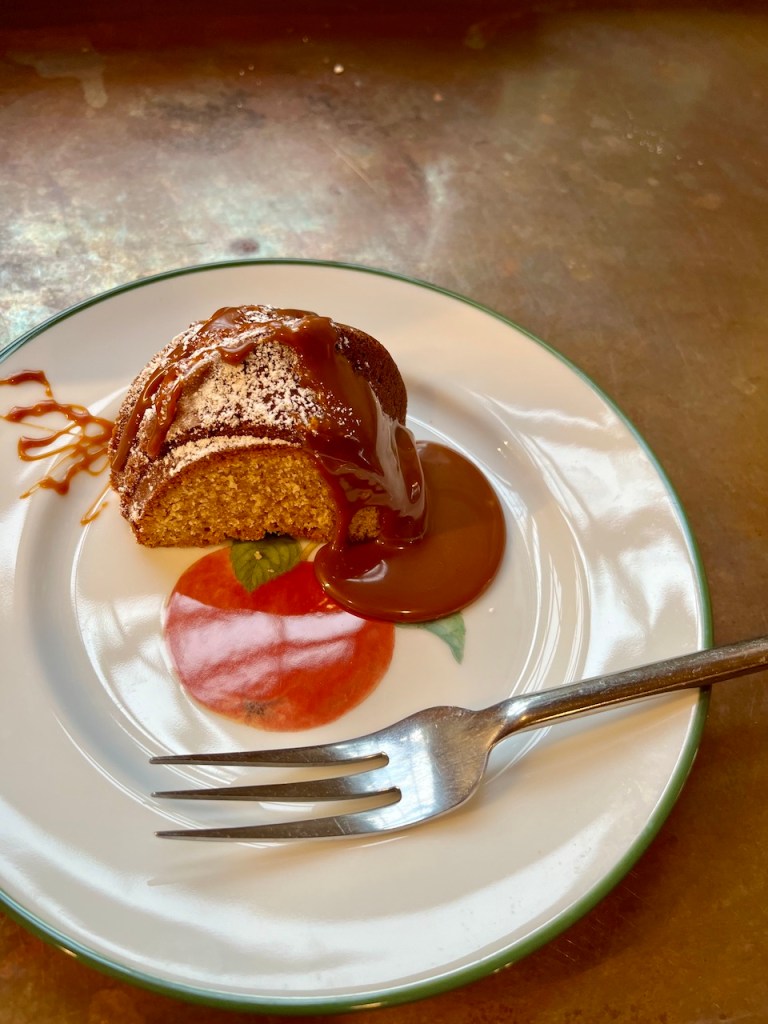 A slice of burnt butter cider cake drizzled with caramel sauce on a white plate with a red apple design, accompanied by a fork.