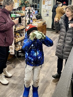 A child wearing a galaxy-themed jacket and Spiderman hat is drinking from a green cup in a cozy indoor setting, while two adults observe in the background.
