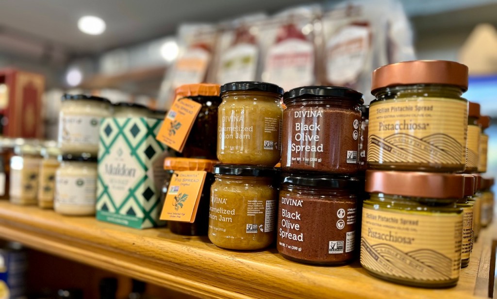 An assortment of gourmet spreads displayed on a wooden shelf, featuring jars of caramelized onion jam, black olive spread, pistachio spread, and more, set against a blurred background of packaged goods.