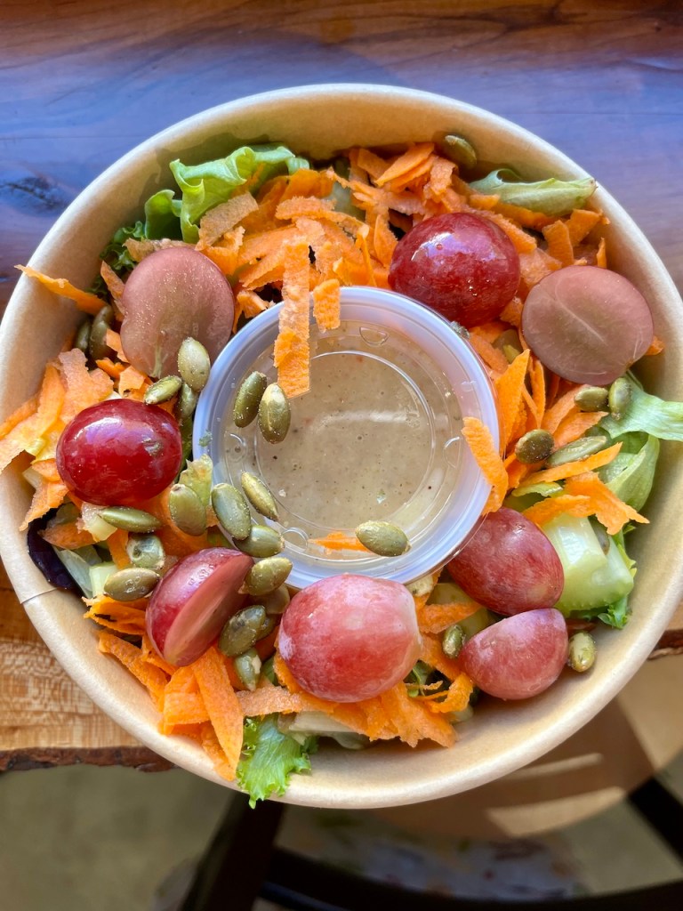 A fresh salad in a round bowl featuring mixed greens, shredded carrots, halved grapes, cucumbers, and pumpkin seeds, accompanied by a small container of dressing.