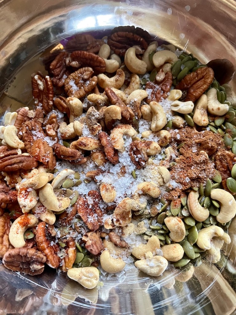 A close-up view of a mixed nut and seed snack blend, including cashews, pecans, pumpkin seeds, and spices, in a metal bowl.