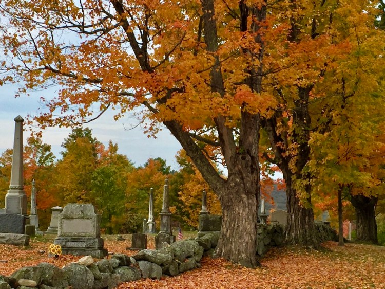 A picturesque cemetery scene featuring autumn foliage with vibrant orange and yellow leaves on trees, gravestones scattered throughout, and a stone wall in the foreground.