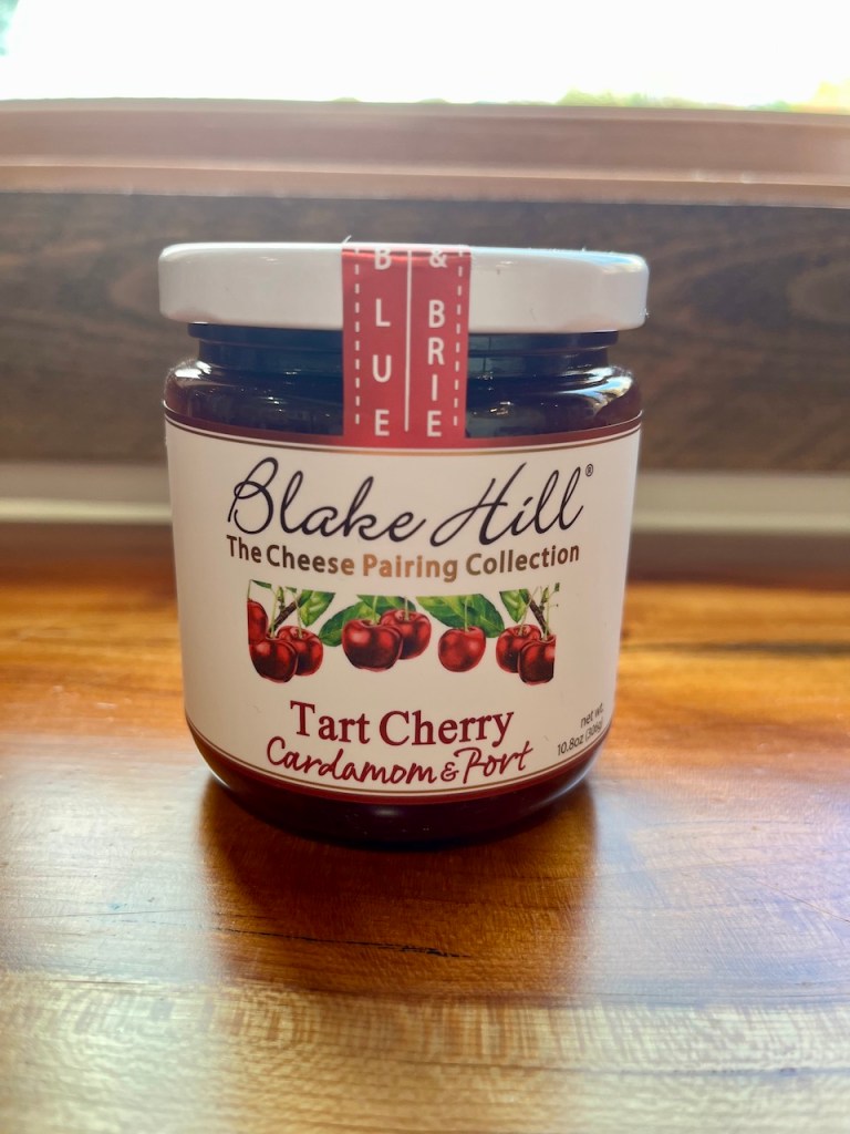 A jar of Blake Hill Tart Cherry jam with Cardamom & Port, placed on a wooden surface, showcasing its label and packaging.