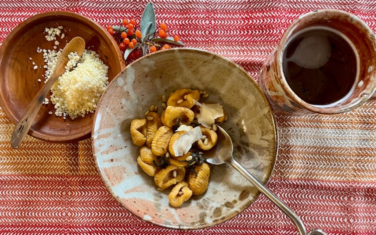 A bowl of pumpkin gnocchi topped with creamy sauce, surrounded by a wooden bowl of grated cheese and a ceramic jug, set on a textured tablecloth.