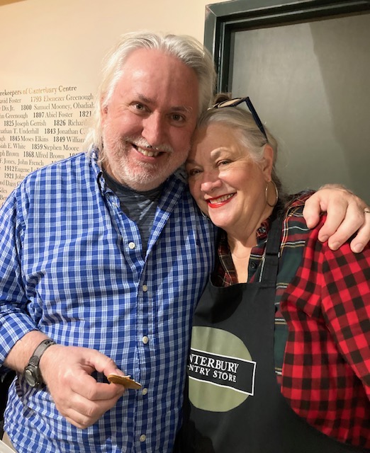 Two smiling individuals posing together in a casual setting, one holding a cookie and wearing a plaid shirt, while the other wears an apron with the Canterbury Country Store logo.